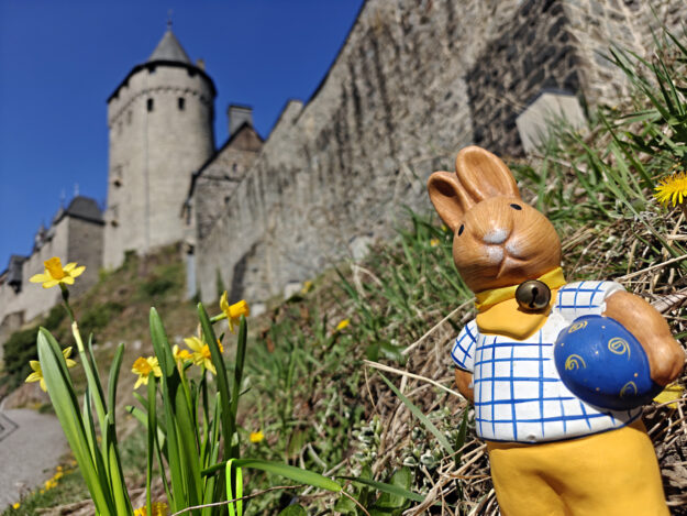 Foto: Märkischer Kreis Eine Osterhasen Figur steht vor den Mauern der Burg Altena, daneben blühen Blumen.