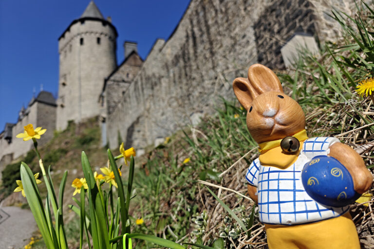 Eine Osterhasen Figur steht vor den Mauern der Burg Altena, daneben blühen Blumen.