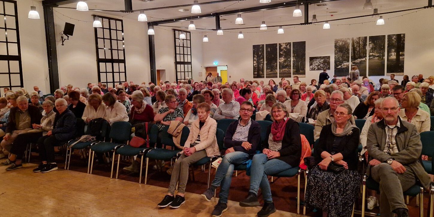 Zuschauer sitzen in Stuhlreihen in einem beleuchteten Saal.