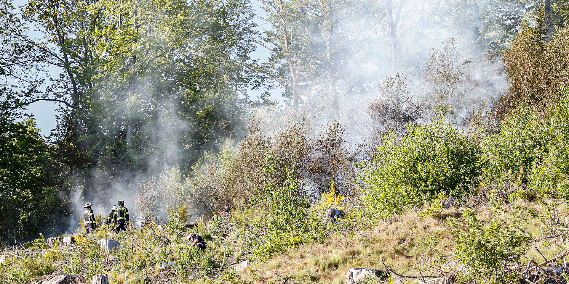 Bei einem Brand an einem steilen Waldgebiet bekämpfen Feuerwehrleute die Flammen. Es steigt Rauch auf.