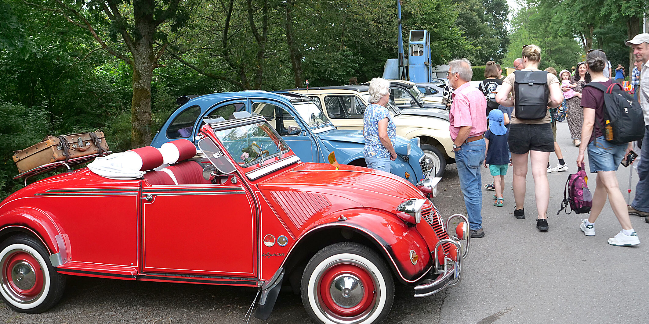 Eine Gruppe von Menschen steht auf einer Straße und betrachtet mehrere klassische Citroën 2CV, auch „Ente“ genannt, bei einem Oldtimertreffen. Im Vordergrund ist ein auffällig restauriertes, rotes Cabrio-Modell mit offenem Verdeck.