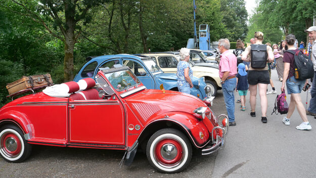 Foto: Märkische Museumseisenbahn Eine Gruppe von Menschen steht auf einer Straße und betrachtet mehrere klassische Citroën 2CV, auch „Ente“ genannt, bei einem Oldtimertreffen. Im Vordergrund ist ein auffällig restauriertes, rotes Cabrio-Modell mit offenem Verdeck.