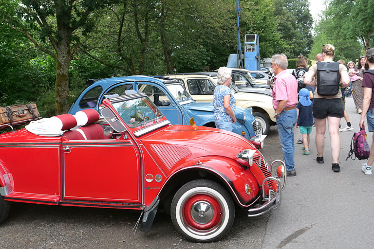 Eine Gruppe von Menschen steht auf einer Straße und betrachtet mehrere klassische Citroën 2CV, auch „Ente“ genannt, bei einem Oldtimertreffen. Im Vordergrund ist ein auffällig restauriertes, rotes Cabrio-Modell mit offenem Verdeck.
