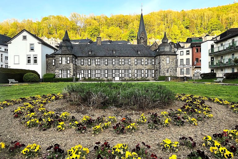 In einer parkähnlichen Gartenanlage blühen dunkelrote und gelbe Stiefmütterchen vor einem burgähnlichen Anwesen.