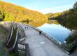 Auf der Staumauer einet Talsperre sind Spaziergänger unterwegs. Auf der Wasseroberfläche spiegeln sich die Bäume der umgebenden Wälder.