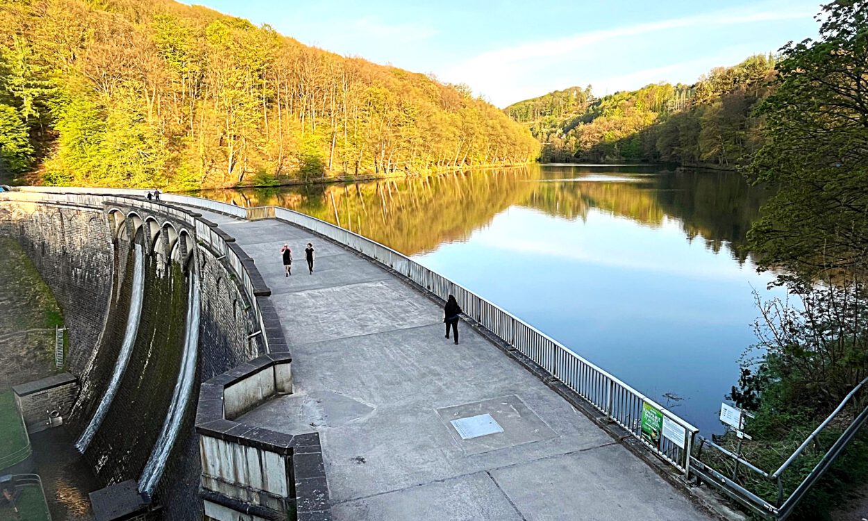 Auf der Staumauer einet Talsperre sind Spaziergänger unterwegs. Auf der Wasseroberfläche spiegeln sich die Bäume der umgebenden Wälder.