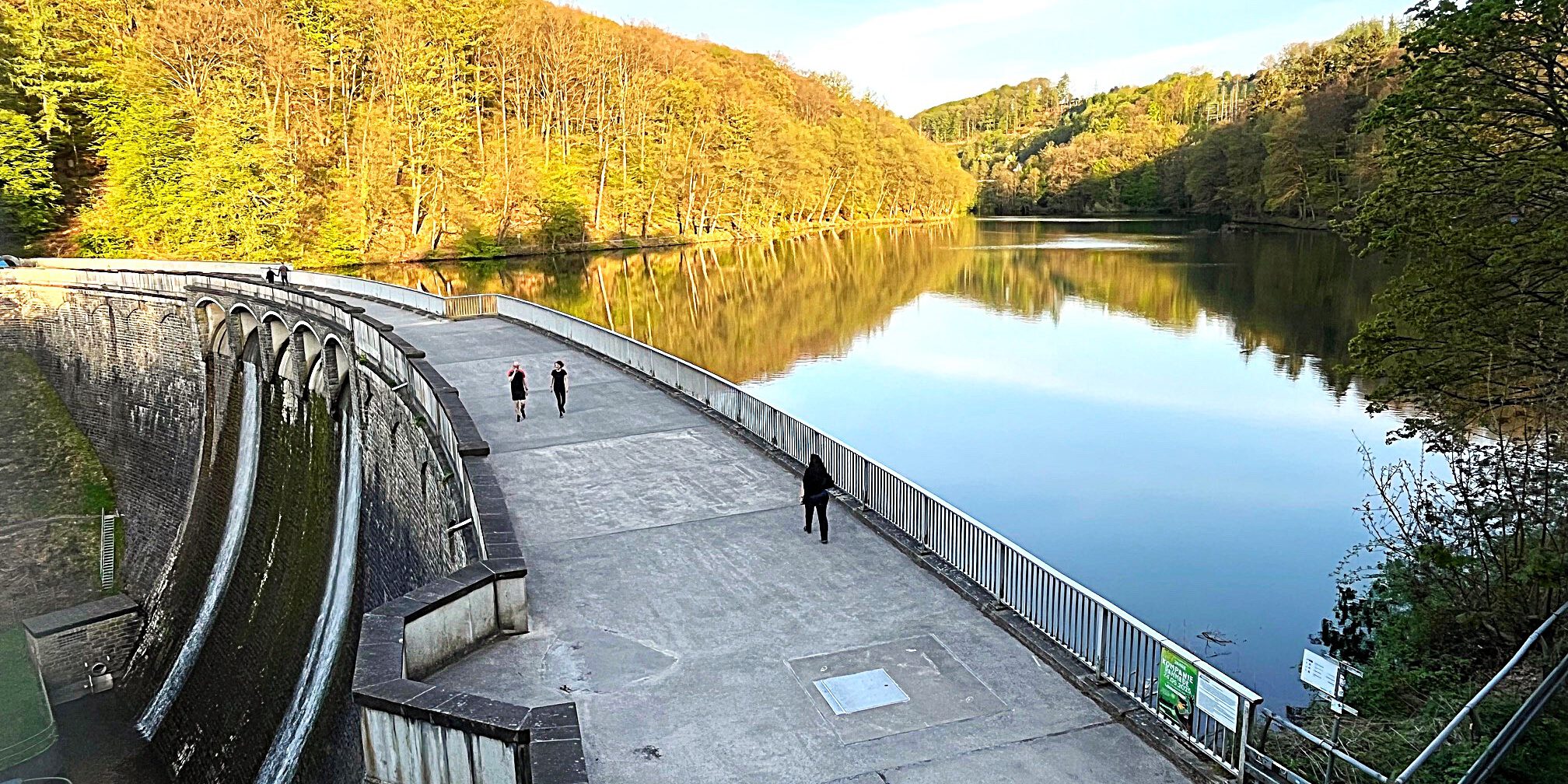 Auf der Staumauer einet Talsperre sind Spaziergänger unterwegs. Auf der Wasseroberfläche spiegeln sich die Bäume der umgebenden Wälder.