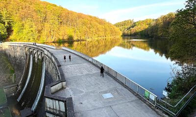 Auf der Staumauer einet Talsperre sind Spaziergänger unterwegs. Auf der Wasseroberfläche spiegeln sich die Bäume der umgebenden Wälder.