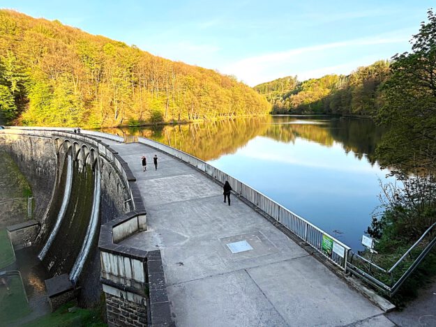 Foto: Carsten Menzel Auf der Staumauer einet Talsperre sind Spaziergänger unterwegs. Auf der Wasseroberfläche spiegeln sich die Bäume der umgebenden Wälder.