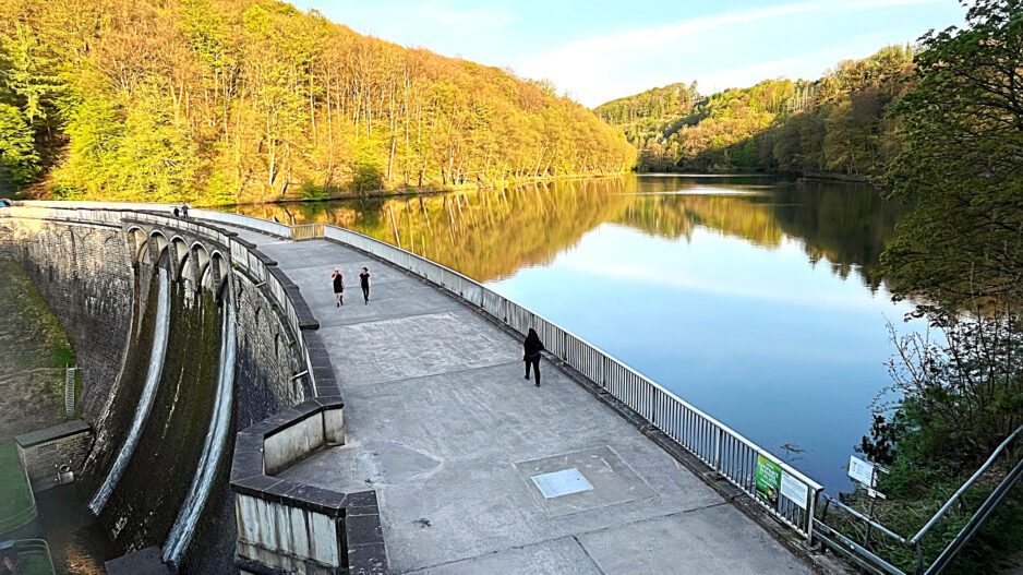 Auf der Staumauer einet Talsperre sind Spaziergänger unterwegs. Auf der Wasseroberfläche spiegeln sich die Bäume der umgebenden Wälder.