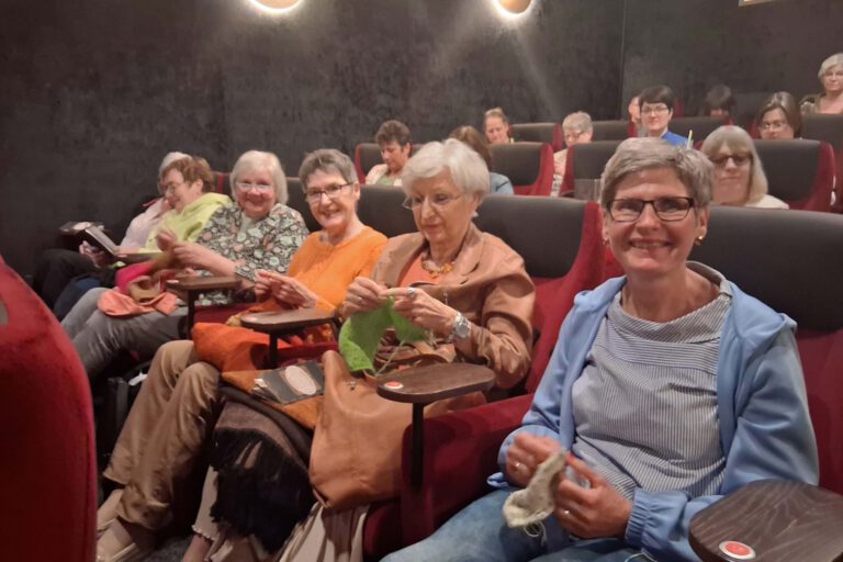 Mehrere Frauen sitzen in einem dunklen Kinosaal und stricken während einer Filmvorstellung.