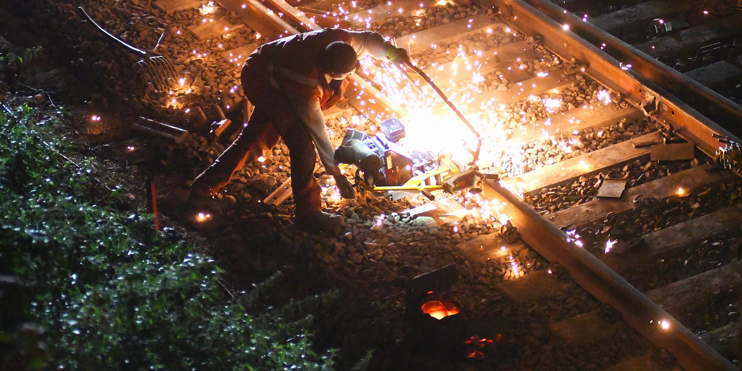 Ein Bauarbeiter füllt an einer Eisenbahnstrecke flüssiges Eisen in eine Lücke in einem Schienenstrang. Es fliegen Funken.