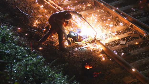 Foto: Carsten Menzel/Archiv Ein Bauarbeiter füllt an einer Eisenbahnstrecke flüssiges Eisen in eine Lücke in einem Schienenstrang. Es fliegen Funken.