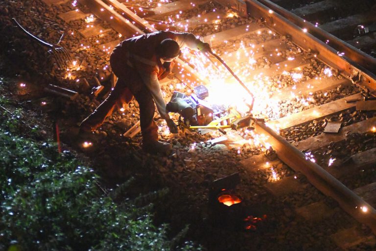 Ein Bauarbeiter füllt an einer Eisenbahnstrecke flüssiges Eisen in eine Lücke in einem Schienenstrang. Es fliegen Funken.
