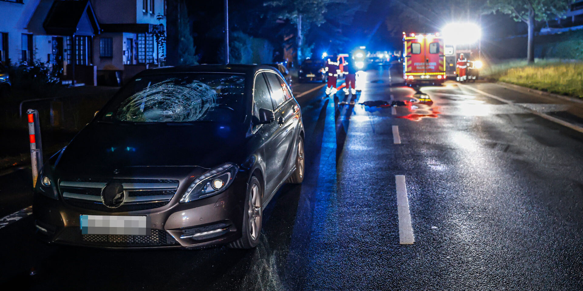 Ein Unfallwagen steht mit beschädigter Windschutzscheibe in der Nacht auf einer Straße. Rettungskräfte arbeiten im Hintergrund.
