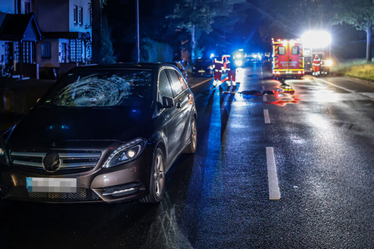Ein Unfallwagen steht mit beschädigter Windschutzscheibe in der Nacht auf einer Straße. Rettungskräfte arbeiten im Hintergrund.