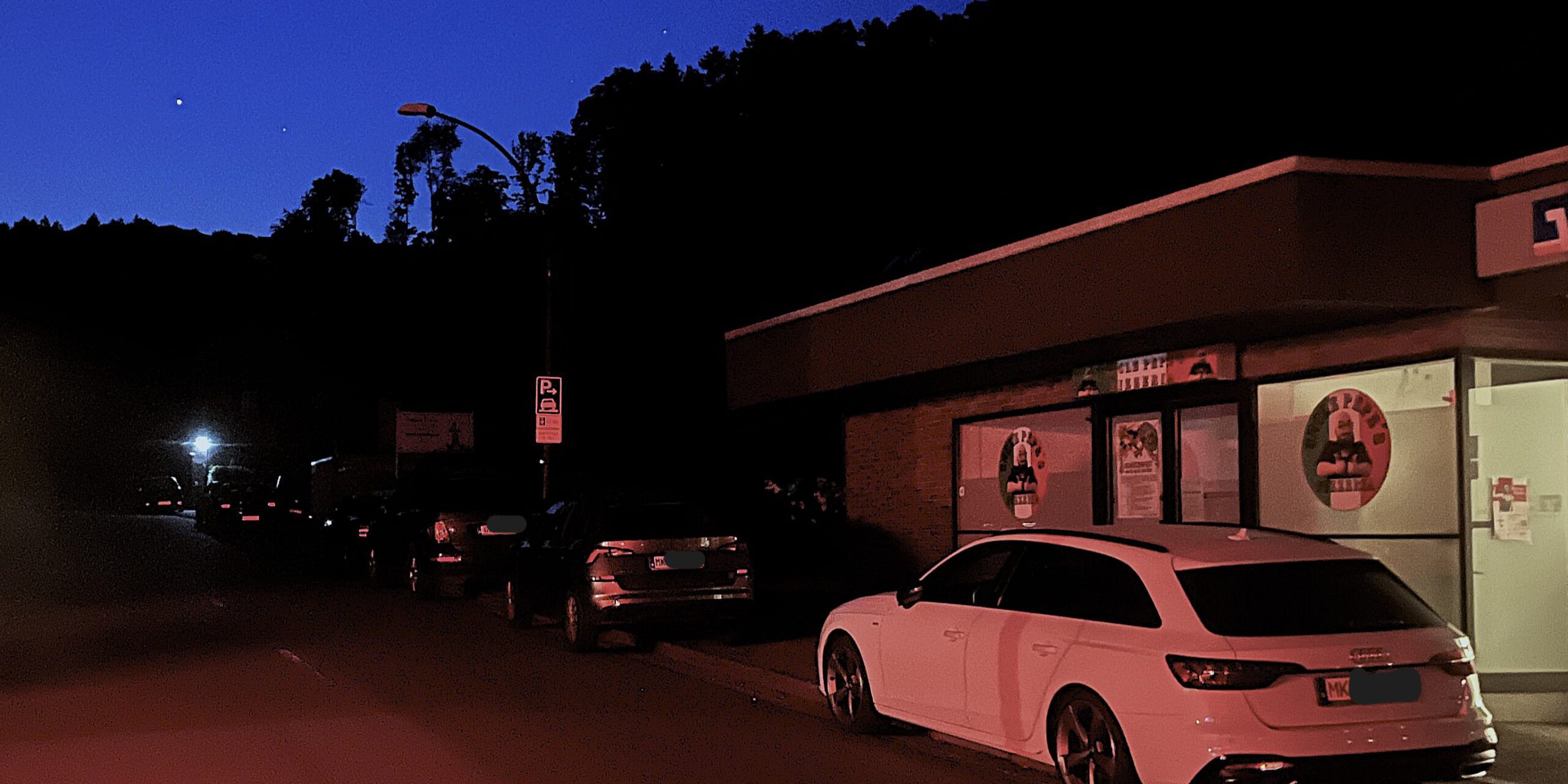 Ein Straßenzug liegt im Dunkel der Nacht. Nur rechts vorne im Bild lässt sich ein weißes Auto durch das Licht einer Schaufensterbeleuchtung erkennen.
