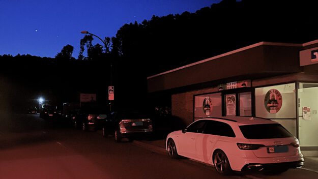 Ein Straßenzug liegt im Dunkel der Nacht. Nur rechts vorne im Bild lässt sich ein weißes Auto durch das Licht einer Schaufensterbeleuchtung erkennen.