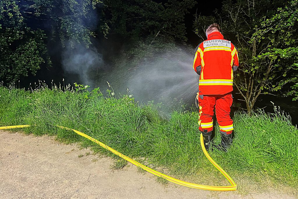 Ein Feuerwehrmann in einer orangen Uniform steht an einer Flussböschung und löscht mit einem C-Rohr Flammen im Unterholz. Rauch steigt auf.