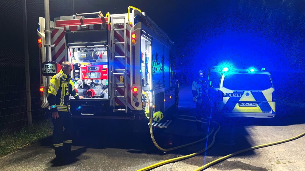 Ein Feuerwehrmann steht am Bedienfeld am Heck eines Feuerwehrautos, an dem ein gelber Schlauch angeschlossen ist. Im Bildhintergrund steht ein Polizeiwagen mit Blaulicht.