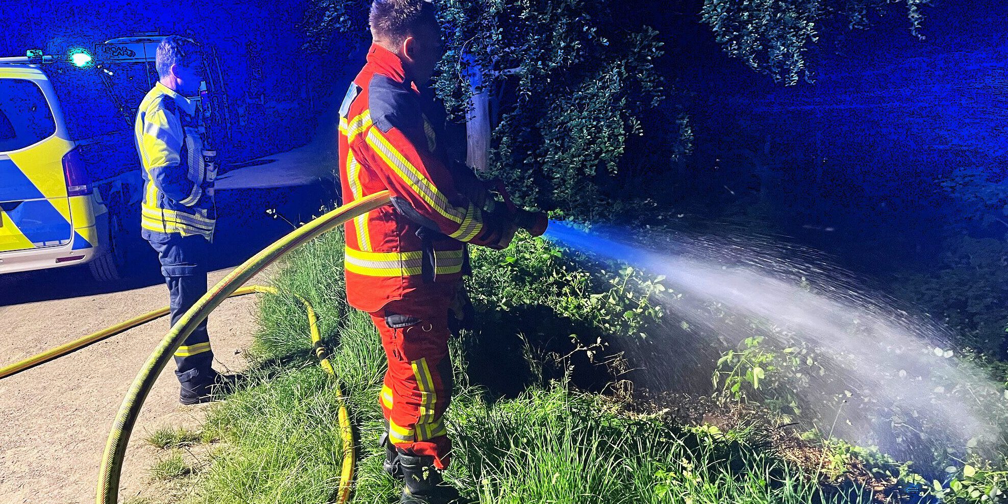 Ein Feuerwehrmann in einer orangen Uniform steht an einer Flussböschung und löscht mit einem C-Rohr Flammen im Unterholz. Im Hintergrund stehen ein weiterer Feuerwehrmann und ein Polizeiwagen mit Blaulicht.