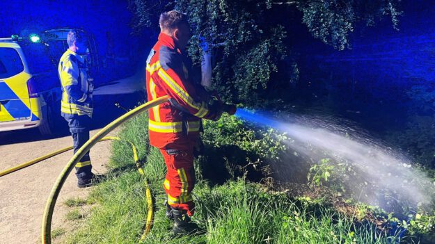 Foto: Carsten Menzel Ein Feuerwehrmann in einer orangen Uniform steht an einer Flussböschung und löscht mit einem C-Rohr Flammen im Unterholz. Im Hintergrund stehen ein weiterer Feuerwehrmann und ein Polizeiwagen mit Blaulicht.