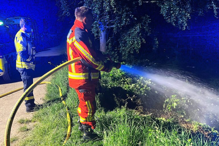 Ein Feuerwehrmann in einer orangen Uniform steht an einer Flussböschung und löscht mit einem C-Rohr Flammen im Unterholz. Im Hintergrund stehen ein weiterer Feuerwehrmann und ein Polizeiwagen mit Blaulicht.