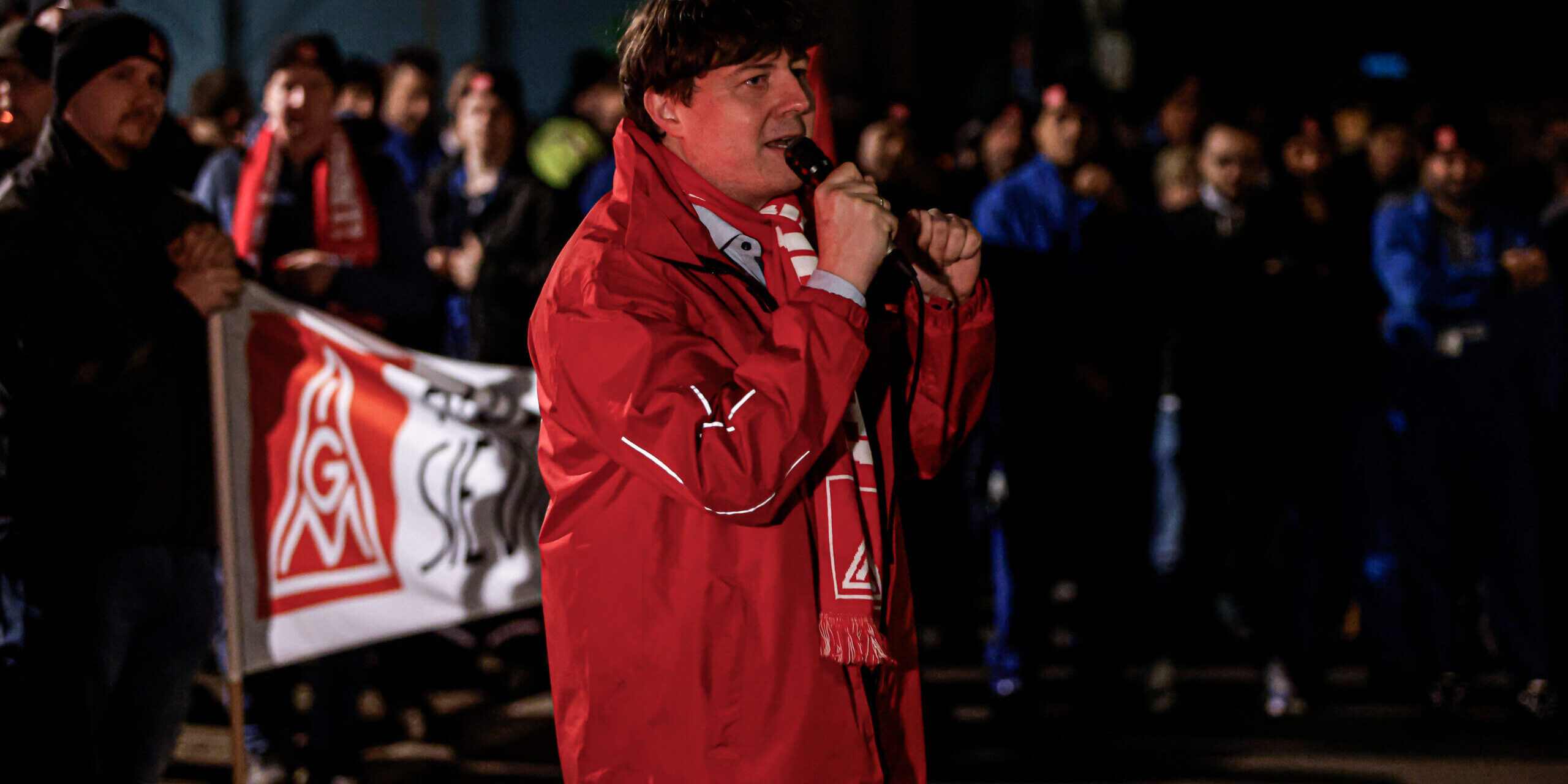 Ein Mann steht in einr roten Jacke und Schal der IG Metall vor einer Gruppe von Arbeitern und spricht in ein Mikrofon.