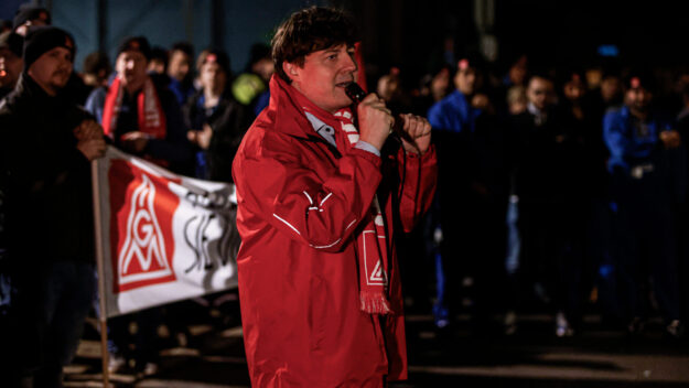Foto: Dennis Echtermann Ein Mann steht in einr roten Jacke und Schal der IG Metall vor einer Gruppe von Arbeitern und spricht in ein Mikrofon.