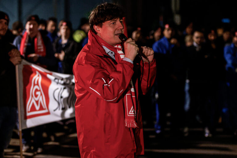 Ein Mann steht in einr roten Jacke und Schal der IG Metall vor einer Gruppe von Arbeitern und spricht in ein Mikrofon.