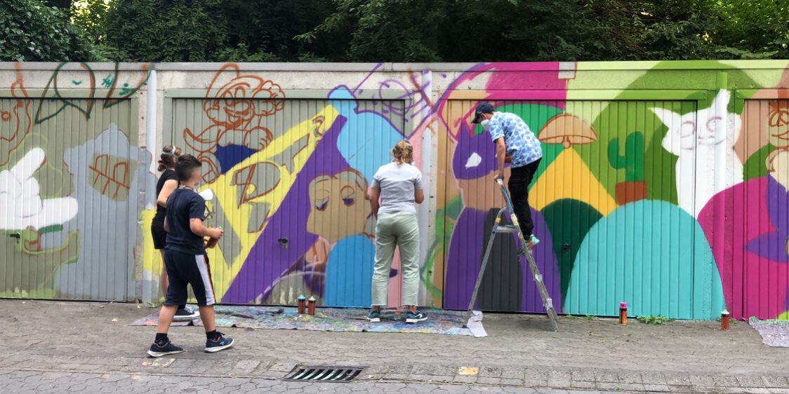 Kinder stehen vor Garagen, die schon zur Hälfte in bunten Farben mit Graffiti besprüht sind. An der anderen Hälfte arbeiten sie gerade.