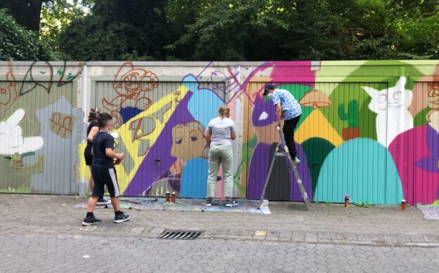 Foto: Stadt Altena Kinder stehen vor Garagen, die schon zur Hälfte in bunten Farben mit Graffiti besprüht sind. An der anderen Hälfte arbeiten sie gerade.