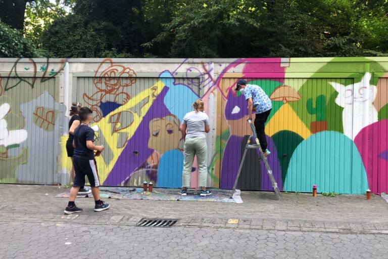 Foto: Stadt Altena Kinder stehen vor Garagen, die schon zur Hälfte in bunten Farben mit Graffiti besprüht sind. An der anderen Hälfte arbeiten sie gerade.