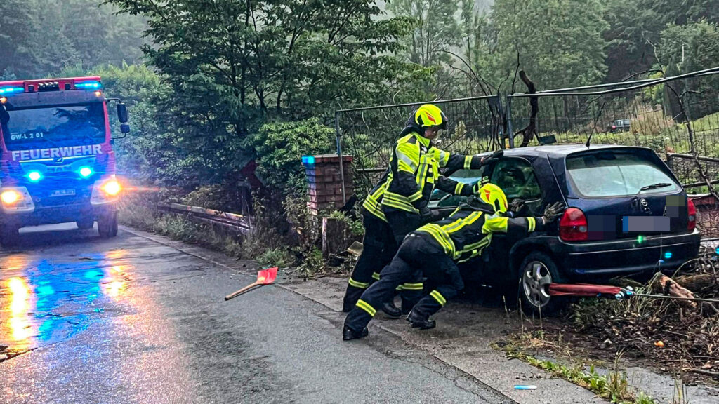 Drei Feuerwehrleute in Uniform drücken gegen einen dunkelblauen Kleinwagen am Straßenrand. Im Hintergrund steht ein Feuerwehrauto mit Blaulicht.