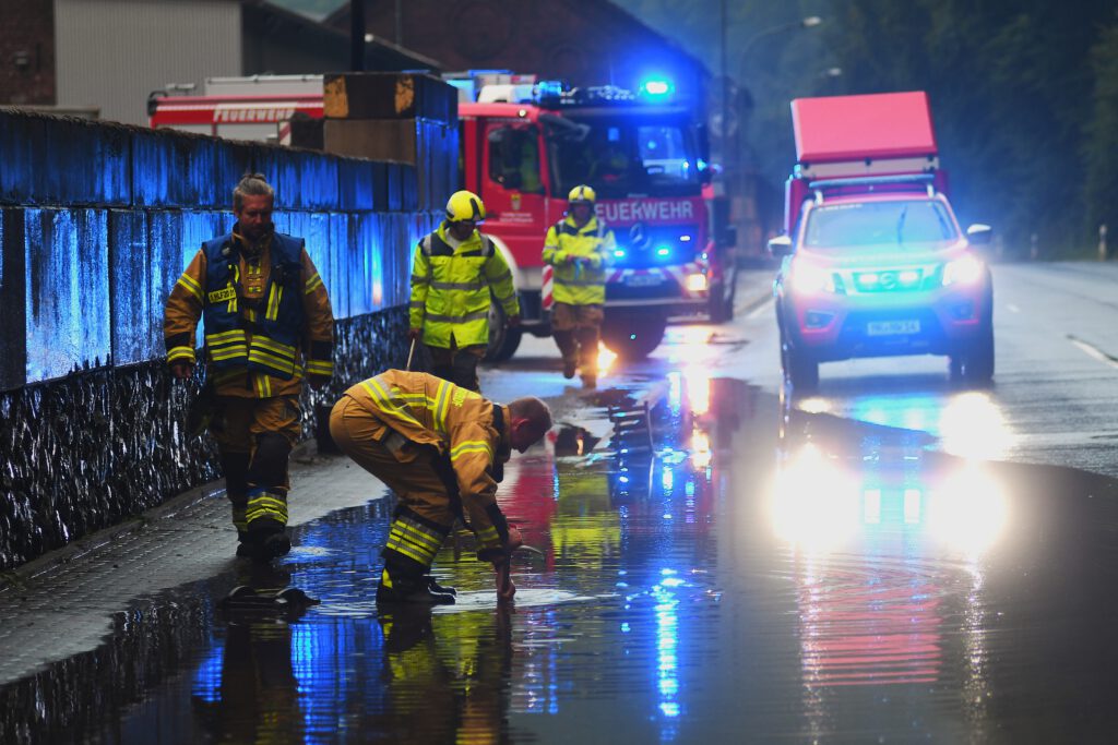 Auf einer Straße sammelt sich Regenwasser. In dem Wasser spiegeln sich zwei Feuerwehrfahrzeuge mit Blaulicht. Vier Feuerwehrleute gehen an der großen Pfütze vorbei.