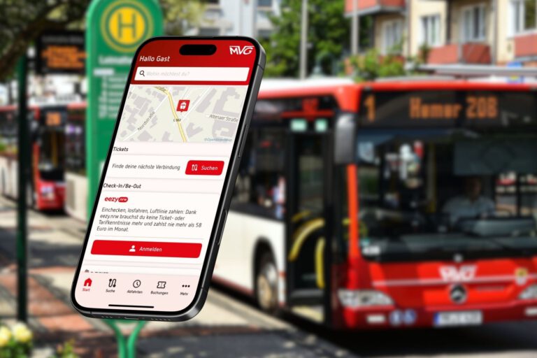 Ein Smartphone, auf dem die neue MVG-App aufgerufen ist vor einem Hintergrund-Foto, das verschwommen einen Bus der MVG an einer Haltestelle zeigt.