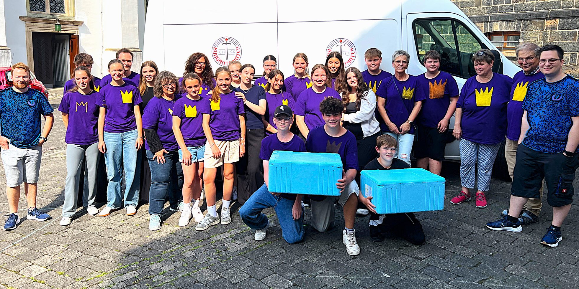 Eine Gruppe junger und älterer Menschen steht auf einem Kirchplatz, drei Jugendliche halten zwei hellblaue Isolierkartons.