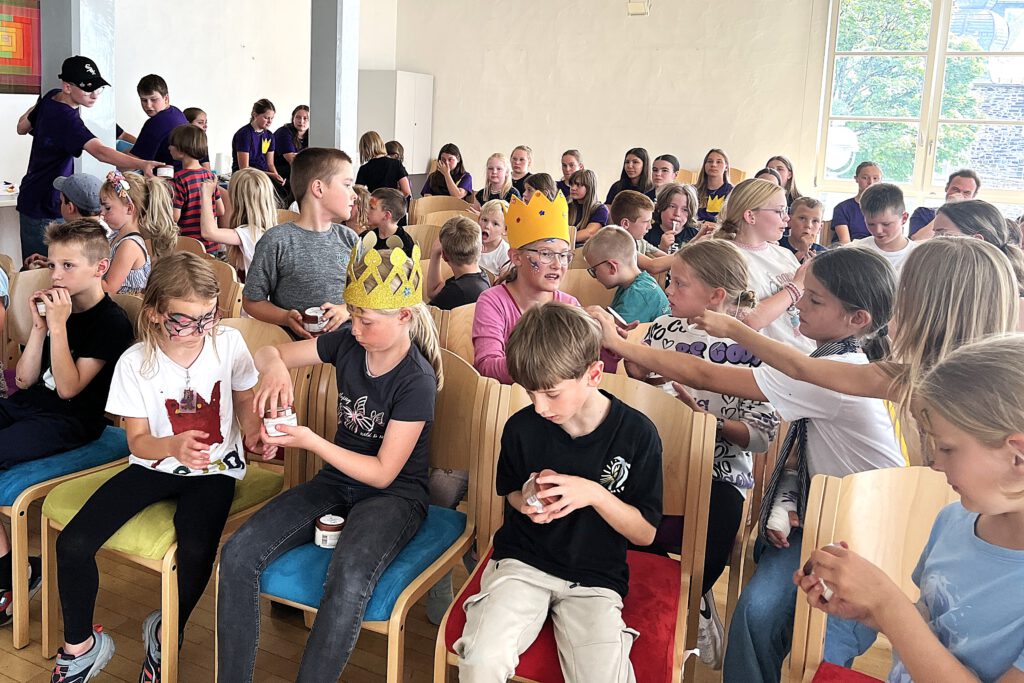 Jugendliche in dunkelblauen T-Shirts verteilen Eisbecher an Kinder in einem Gemeindesaal.
