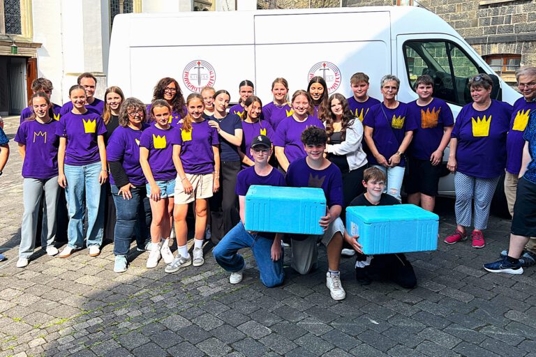 Foto: Ev. Kirchengemeinde Eine Gruppe junger und älterer Menschen steht auf einem Kirchplatz, drei Jugendliche halten zwei hellblaue Isolierkartons.