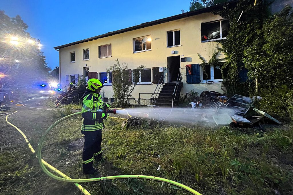 Ein Feuerwehrmann löscht verbrannte Einrichtungsgegenstände, die vor einem Wohnhaus liegen.