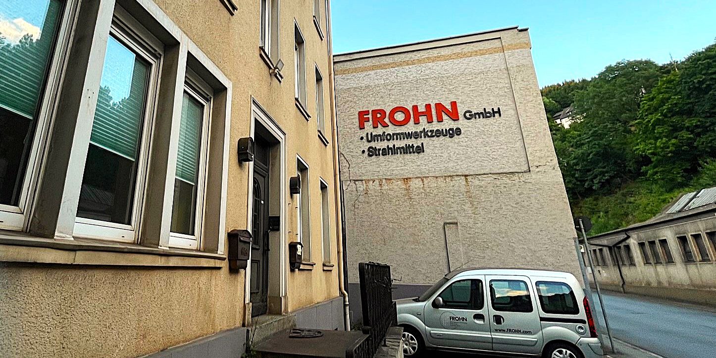 Vor einem beigen Industriegebäude parkt ein silbernes Firmenfahrzeug. Im Hintergrund verläuft eine Straße.