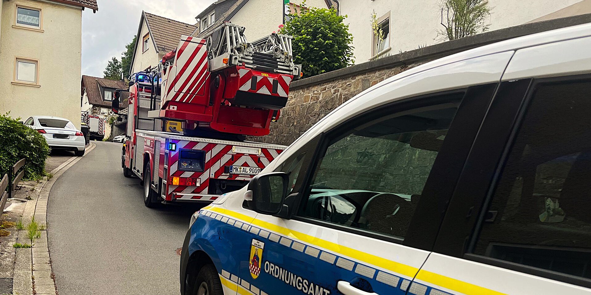Ein Drehleiterfahrzeug der Feuerwehr und ein Wagen des städtischen Ordnungsamts stehen hintereinander in einer engen, bergauf führenden Straße, rechts und links dichte Bebauung durch Wohnhäuser.