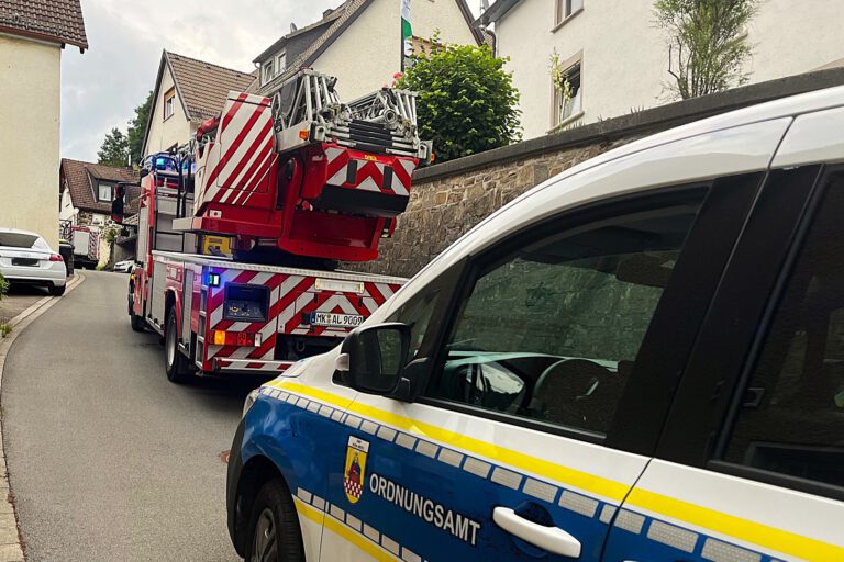 Ein Drehleiterfahrzeug der Feuerwehr und ein Wagen des städtischen Ordnungsamts stehen hintereinander in einer engen, bergauf führenden Straße, rechts und links dichte Bebauung durch Wohnhäuser.