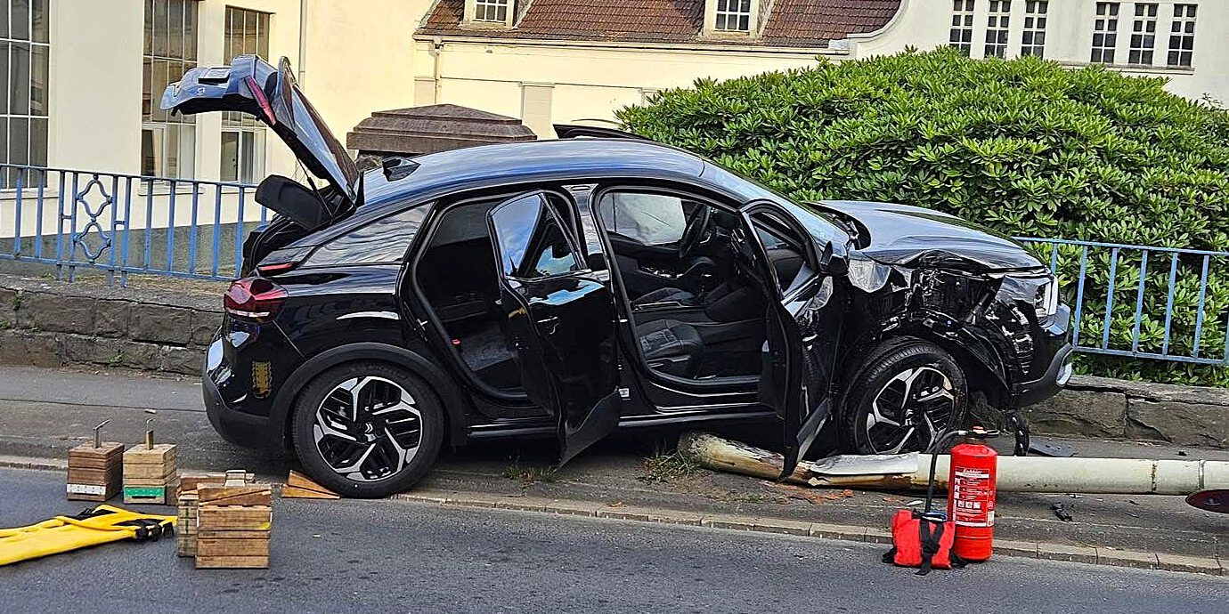 Ein schwarzes Auto steht auf einer umgefahrenen Straßenlaterne, der rechte Kotflügel ist verbeult. Die Front des Autos hängt auf einer kniehohen Mauer am Straßenrand.