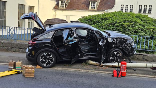 Ein schwarzes Auto steht auf einer umgefahrenen Straßenlaterne, der rechte Kotflügel ist verbeult. Die Front des Autos hängt auf einer kniehohen Mauer am Straßenrand.