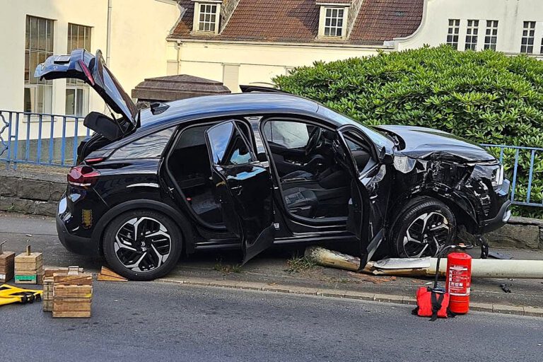 Ein schwarzes Auto steht auf einer umgefahrenen Straßenlaterne, der rechte Kotflügel ist verbeult. Die Front des Autos hängt auf einer kniehohen Mauer am Straßenrand.