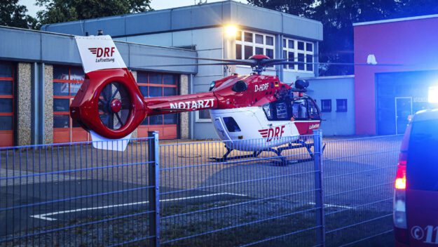Ein rot-weißer Rettungshubschrauber steht in der Morgendämmerung auf einem Platz vor einem Feuerwehrgerätehaus.