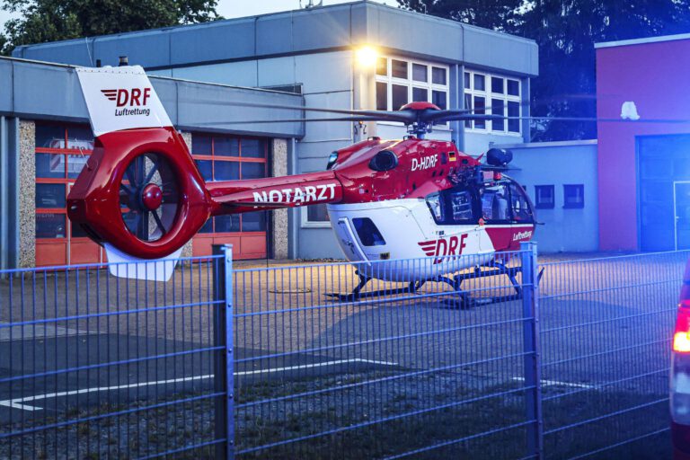 Ein rot-weißer Rettungshubschrauber steht in der Morgendämmerung auf einem Platz vor einem Feuerwehrgerätehaus.