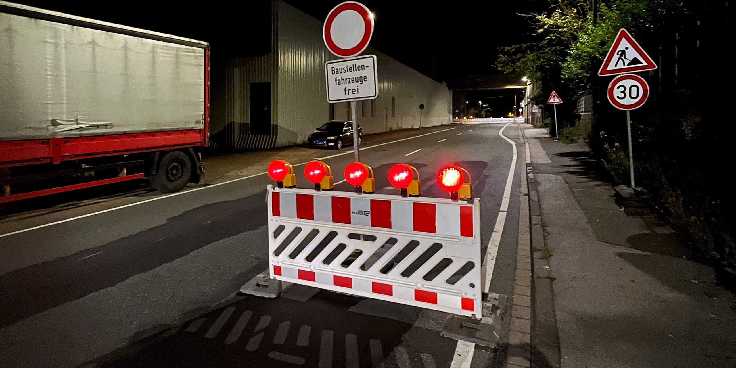 Auf einer nächtlichen Straße stehen rot-weiße Absperrbaken, rote Blinklichter und ein Schild "Durchfahrt verboten - Baustellenfahrzeuge frei"