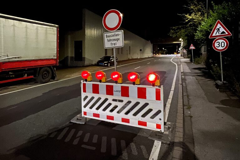 Auf einer nächtlichen Straße stehen rot-weiße Absperrbaken, rote Blinklichter und ein Schild "Durchfahrt verboten - Baustellenfahrzeuge frei"